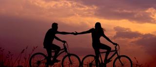 A couple reaching out to each other while riding bicycles at sunset