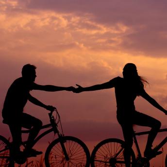 A couple reaching out to each other while riding bicycles at sunset