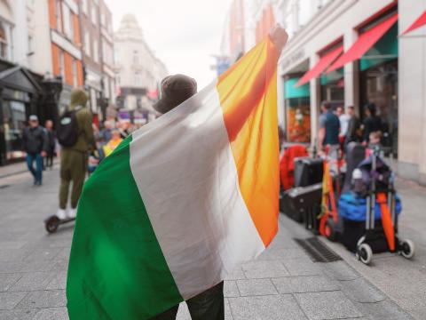 A person on the street, seen from behind, holding up the flag of Ireland