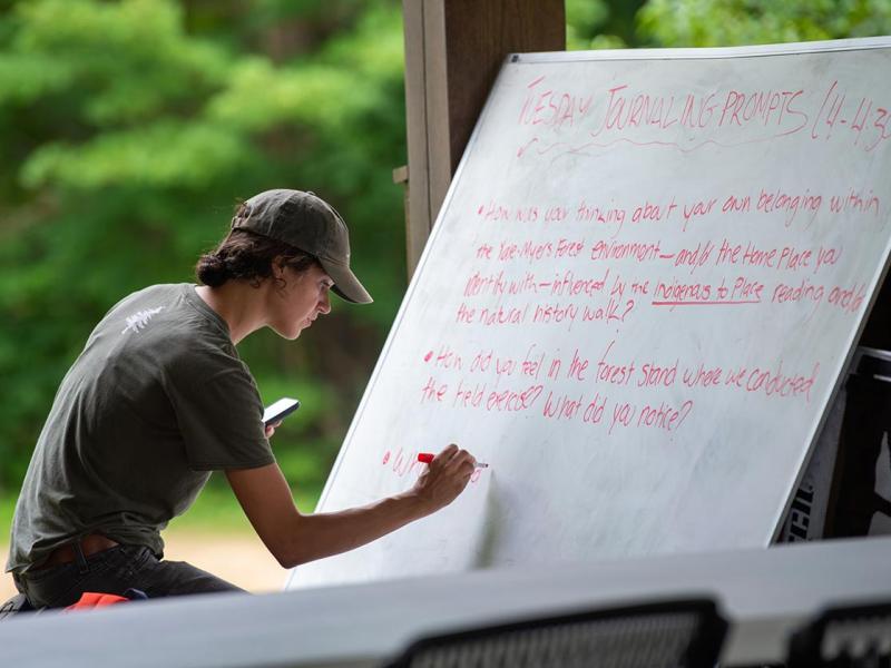 A teaching assistant writing on a large whiteboard in an outdoor auditorium