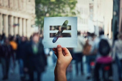 photograph of hand holding up a not-equal sign in public, with out-of-focus passers-by in the background